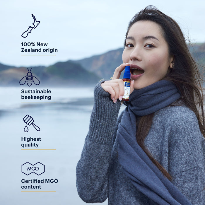 A person stands outdoors by the sea, wearing a gray sweater and scarf, holding a product near their mouth. Text reads: "100% New Zealand origin, Sustainable beekeeping, Highest quality, Certified MGO content.
