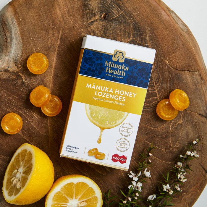 A pack of Manuka Healths Mānuka Honey Lozenges (MGO 400+) sits on wood, surrounded by three lozenges, half a lemon, a wedge, and white flowers with green leaves. The packaging features vibrant blue and yellow branding.