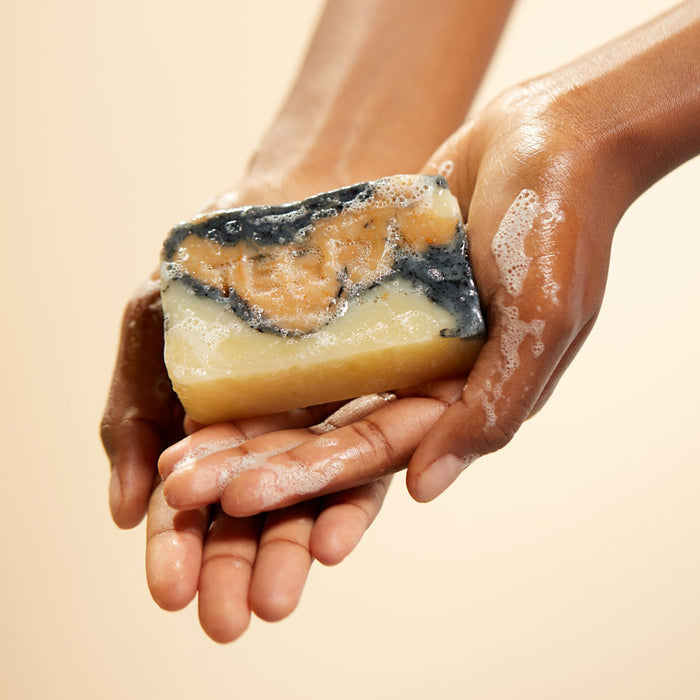 Hands holding a Marble Activated Charcoal Facial Bar with Brazilian Yellow Clay by Terra Beauty Bars, showcasing black, beige, and cream swirls against a neutral background. Soap bubbles accentuate its role as an essential natural facial cleanser.