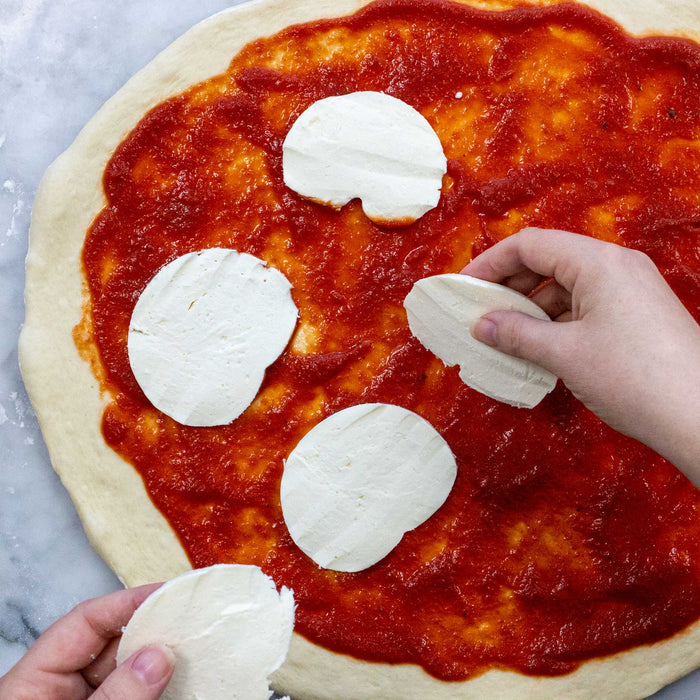 Hands skillfully arrange fresh mozzarella slices on pizza dough coated with rich tomato sauce from the FarmSteady Margherita Pizza Making Kit, crafting a classic pizza on a cool marble surface.