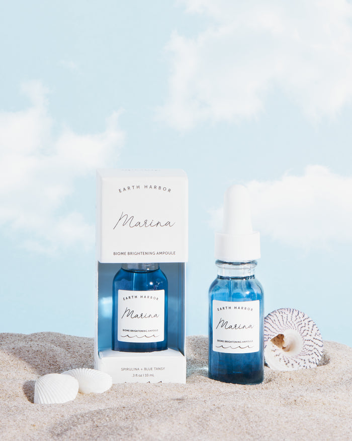 A dropper bottle labeled MARINA Biome Brightening Ampoule from Earth Harbor rests on sandy shores under a bright sky with fluffy clouds. Surrounded by seashells, it captures coastal charm while adaptogenic oils offer microbiome brightening benefits.