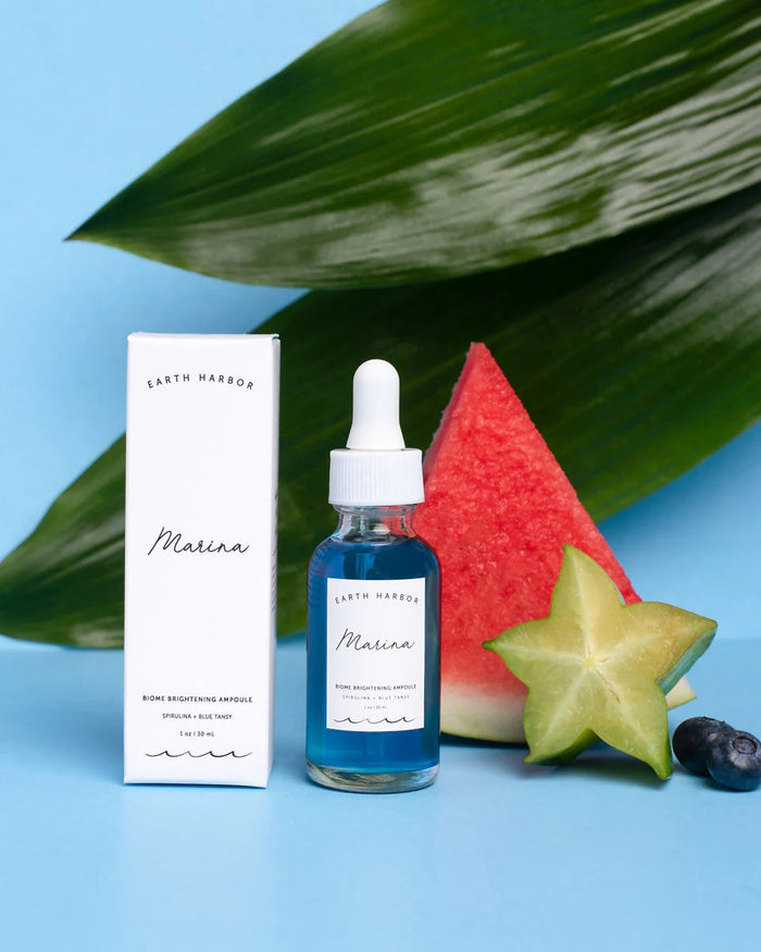 A blue serum bottle labeled MARINA Biome Brightening Ampoule by Earth Harbor stands beside its box, with watermelon, starfruit, blueberries, and green leaves on a vibrant surface. Enriched with ocean-blue botanicals, it nurtures your skin microbiome.