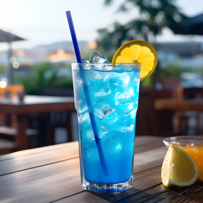A tall glass of Kuli Kuli Foods’ Marine Glow Organic Luminizing Blend, a bright blue iced drink with marine minerals, lemon slice, and blue straw, sits on a wooden table outdoors amid blurred greenery and tables.