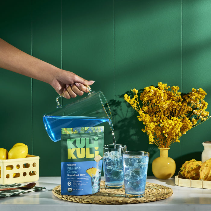 A hand pours vibrant blue Marine Glow Organic Luminizing Blend from Kuli Kuli Foods over ice into glasses near its pouch. Lemons and yellow flowers decorate the table, set against a green wall.