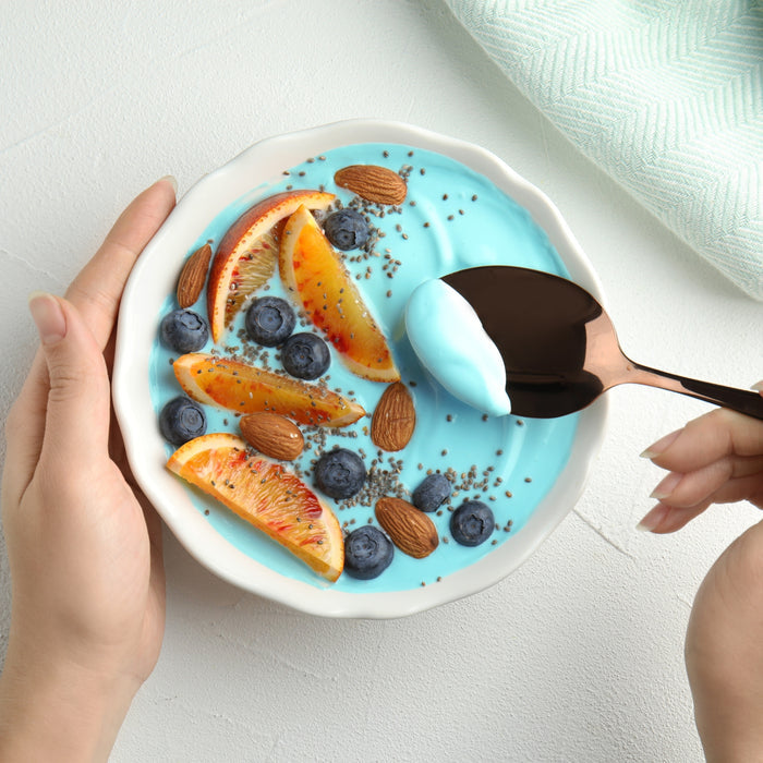 A bowl of blue yogurt made with Kuli Kuli Foods’ Marine Glow Organic Luminizing Blend is topped with orange slices, blueberries, almonds, and chia seeds. A hand holds the bowl while another lifts a spoonful above a white surface and green cloth.