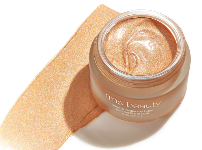 An open jar of RMS Beautys Master Radiance Base reveals a shimmering gold cream, offering a luminous swatch beside it. Proudly displaying the brand, this highlighter features organic ingredients for a radiant look.