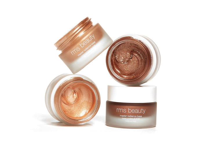 Three open jars of RMS Beautys Master Radiance Base, containing a shimmery highlighting cream in radiant bronze and gold shades, are arranged diagonally. The white caps, labeled clearly, sit beside the small glass containers.