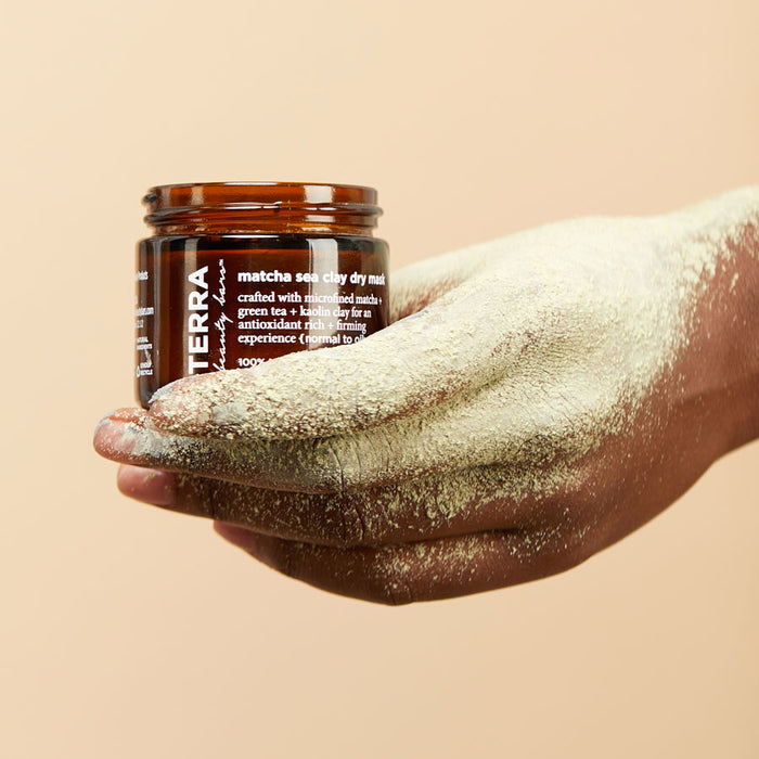 A hand dusted in green powder holds a small brown jar of Terra Beauty Bars Matcha Sea Dry Facial Clay Mask, with white text, against a beige backdrop. The clay masks antioxidant-rich formula is crafted using vegan ingredients.