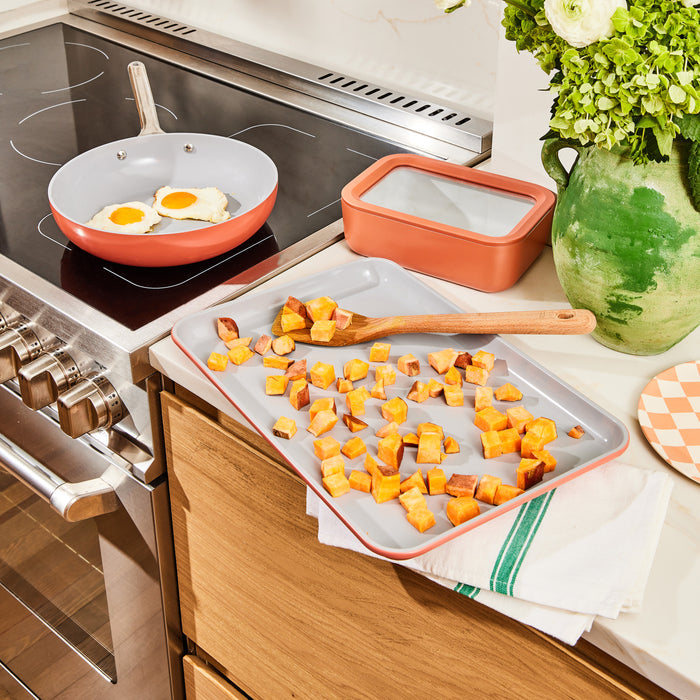 In a kitchen scene, two fried eggs sizzle in a pan on the stove. A Caraway Medium Ceramic Baking Sheet showcases roasted sweet potato cubes, promoting healthy baking, while a wooden spatula rests nearby. A green vase with white flowers stands gracefully against the checkered surface in the background.