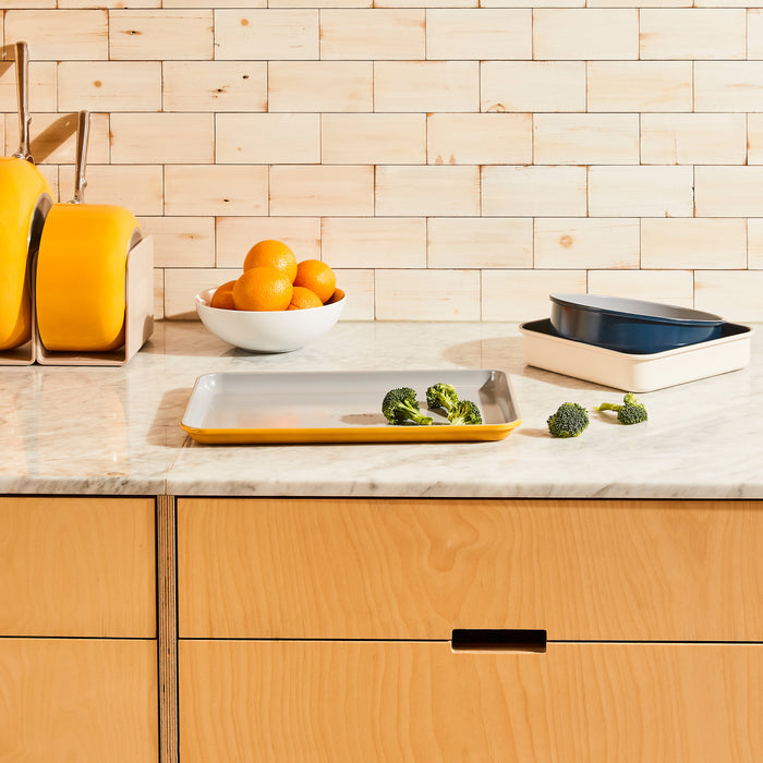 The marble kitchen counter displays a tray of broccoli, a bowl of oranges, and yellow canisters next to stacked blue and white dishes. The backsplash features light wooden tiles. Nearby, a Caraway medium ceramic baking sheet ensures healthy baking adventures.