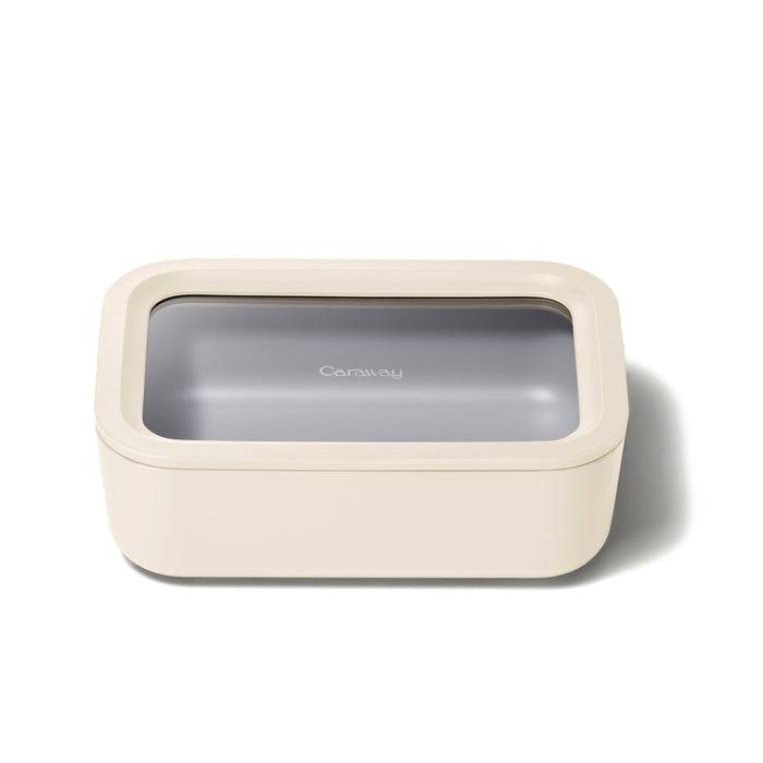 The Medium Glass Food Container by Caraway is beige and rectangular, with a transparent lid displaying the brand name. Made from ceramic-coated glass, it features smooth edges and a minimalist design. Air Release technology ensures its non-toxic storage solution.