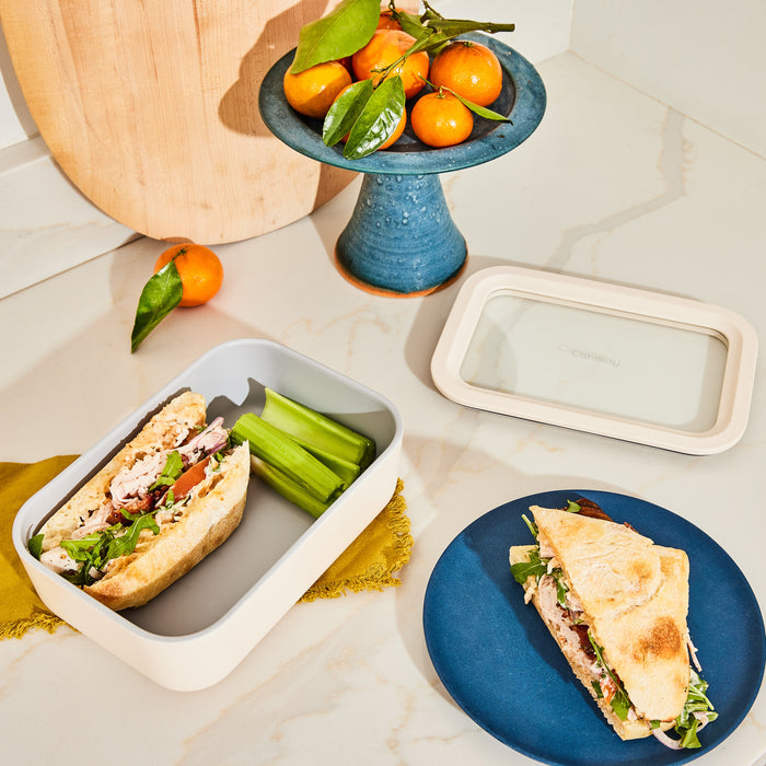 A sandwich with lettuce and meat rests in a Caraway Medium Glass Food Container next to celery sticks on marble. Nearby, a similar sandwich is on a blue ceramic-coated glass plate. A blue pedestal displays fresh oranges with leaves, with a wooden board in the background.