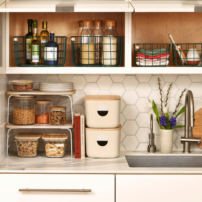 A kitchen counter with neatly organized Open Spaces Medium Storage Bins - Wooden Lids holding spice jars, cooking oils, and wire baskets. Multi-purpose containers and jars with dry goods are complemented by a vase of purple flowers. The backsplash features hexagonal tiles.
