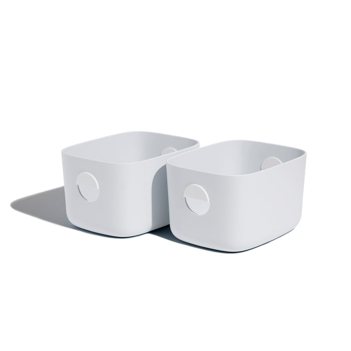 Two Open Spaces Medium Storage Bins, featuring rounded corners and circular handles, are neatly placed side by side on a shelf against a white background. They cast soft shadows to the left, adding an elegant touch of multi-purpose storage.