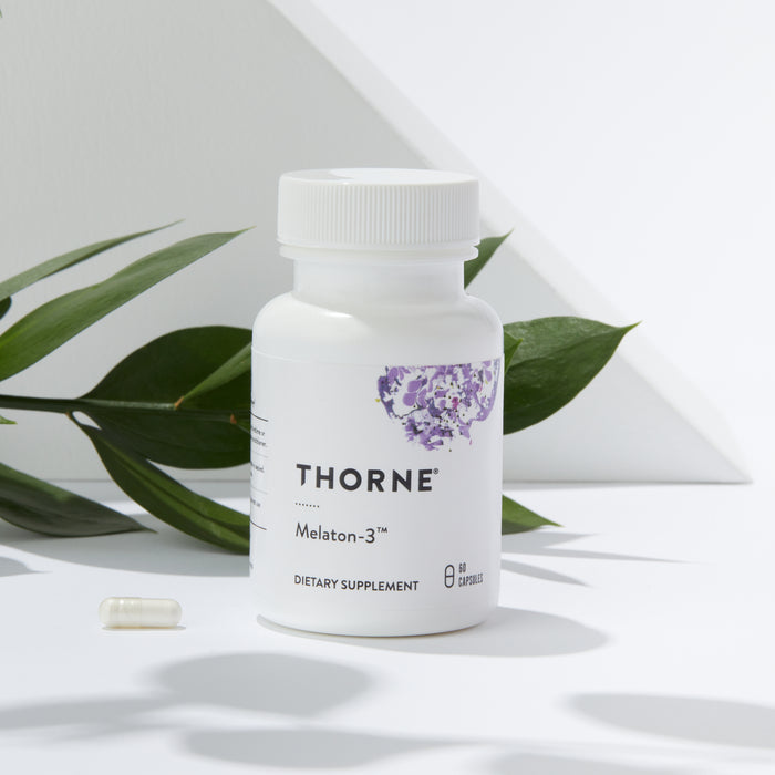 A Thorne Melaton-3 bottle sits by a capsule on a minimalist backdrop of green leaves and geometric shadows, offering restful sleep support.