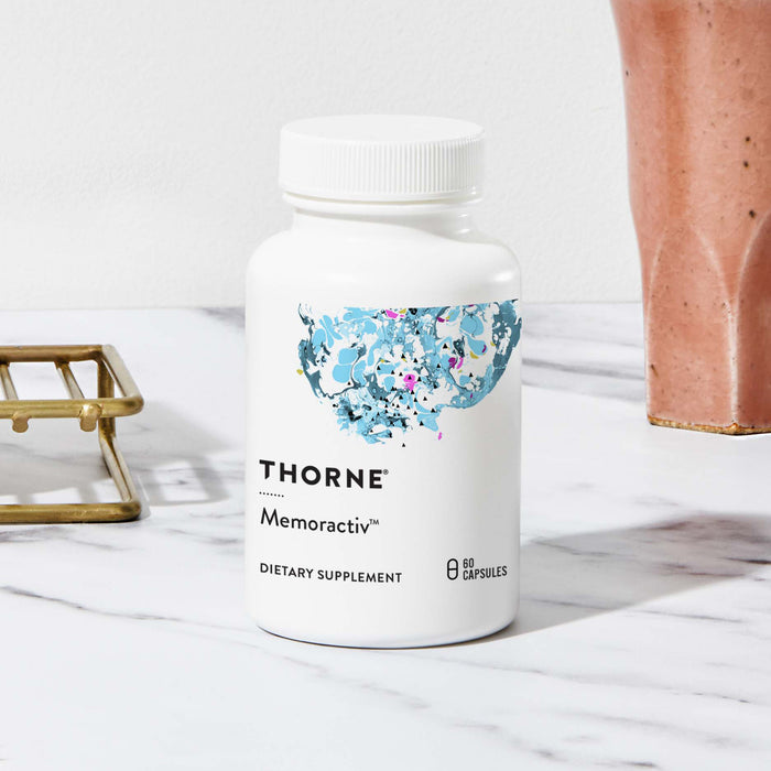 A bottle of Thornes Memoractiv™ cognitive supplement sits elegantly on a marble surface, featuring abstract blue and pink artwork, complemented by a bronze holder and coral-colored pot.