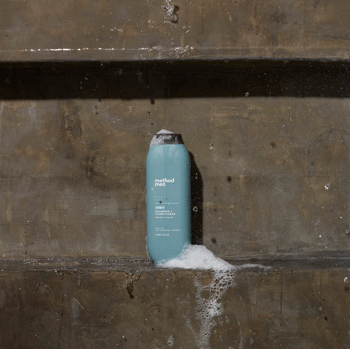 A teal bottle of method Mens 2-in-1 Shampoo + Conditioner sits on the wet concrete amidst suds and bubbles, with water droplets glistening on the surface, hinting at the refreshing menthol aroma inside.