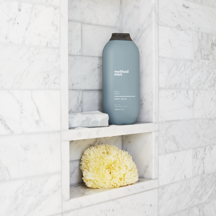 A gray bottle of method Mens Body Wash featuring naturally derived soaps sits on a marble shower shelf, alongside a yellow sponge resting below against pristine white marble tiles.