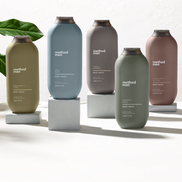 Five bottles of method Mens Body Wash, each in a different color representing scents like Sea + Surf and Cedar + Cypress, are displayed on white pedestals against a neutral background. A green leaf peeks in from the left, emphasizing these naturally derived personal-care products.