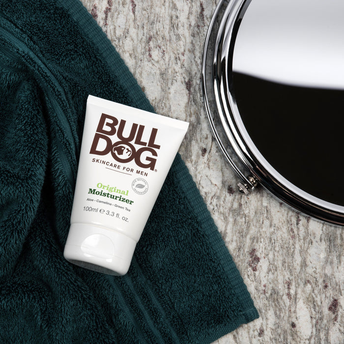 A tube of Bulldog Mens Original Moisturizer, featuring aloe vera and camelina oil for male skin hydration, sits on a dark green towel next to a partially visible metal mirror on a marbled surface.