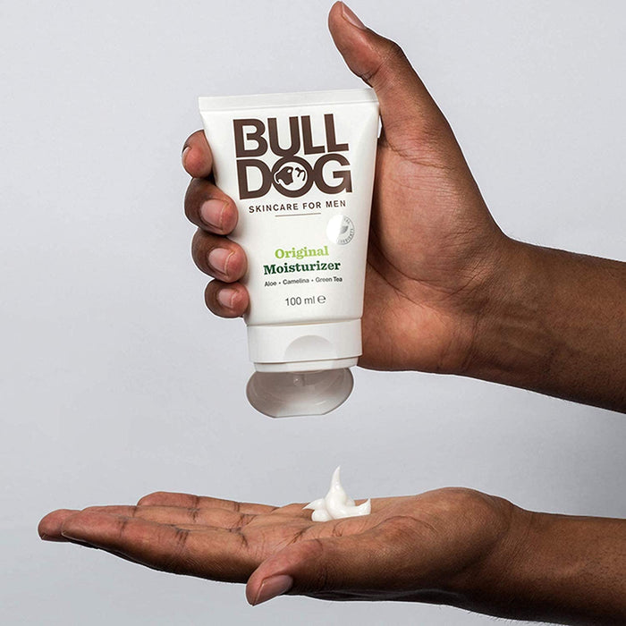 A person holds Bulldog Mens Original Moisturizer, squeezing a small amount into their hand. Infused with aloe vera and camelina oil, it promises optimal hydration for mens skin. The background is plain and neutral.