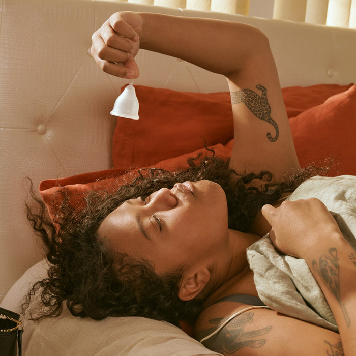 A curly-haired person reclines on a couch, holding a Cora Menstrual Cup. Their tattooed arm is raised while they relax under a soft blanket. Eco-friendly period care essentials and orange cushions provide comfort around them.