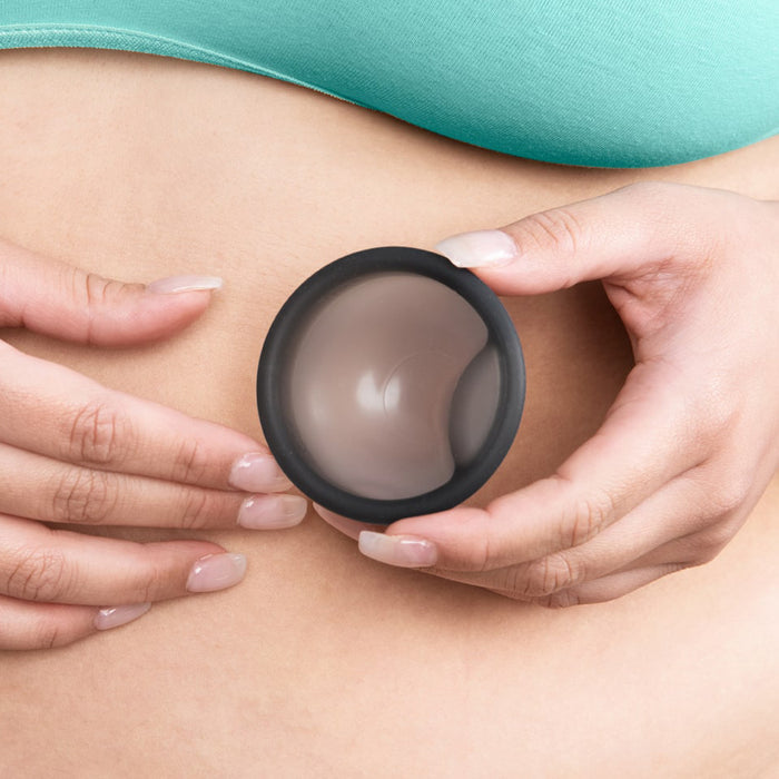 A person holds a contraceptive diaphragm against their abdomen, resembling the ease of a Cora Menstrual Disc with its soft silicone membrane and flexible rim for leak-free protection. Their teal garment enhances the scene, while manicured nails add elegance.