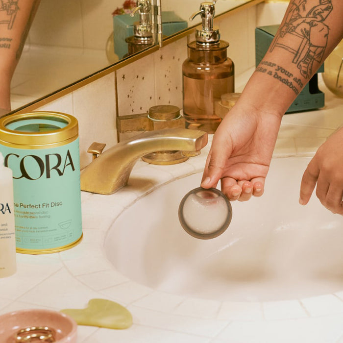 A tattooed person holds a Cora Menstrual Disc, famous for its leak-proof protection, over a sink. Beside them on the tiled counter is a container labeled Cora, along with assorted bathroom items and a green jade facial roller.