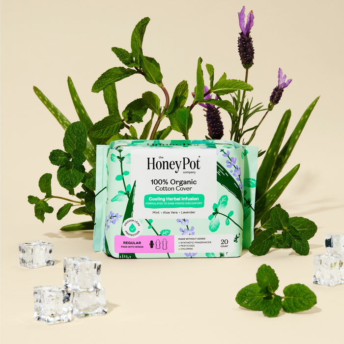 A box of The Honeypot Company 100% Organic Cotton Cover Regular Menstrual Pads - Herbal-Infused is surrounded by mint leaves, aloe vera, lavender flowers, and ice cubes on a beige background.