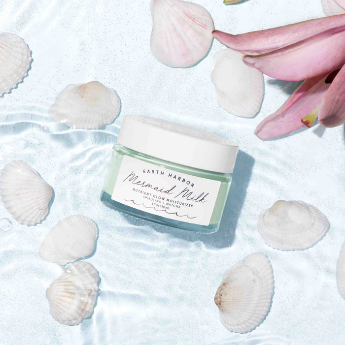 A jar of Earth Harbors MERMAID MILK Nutrient Glow Moisturizer floats on rippling water with pink petals and seashells. This light green, vegan moisturizer features superfoods Spirulina & Matcha and is topped with a white lid.