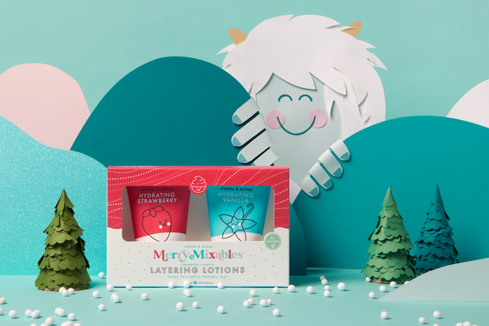 A festive box of Dabble & Dollops Merry Mixables Layering Lotions with hydrating strawberry and vanilla creams is set against a whimsical winter scene featuring a smiling paper yeti among teal mountains and pine trees, with snowflakes accentuating this holiday-themed gift set.
