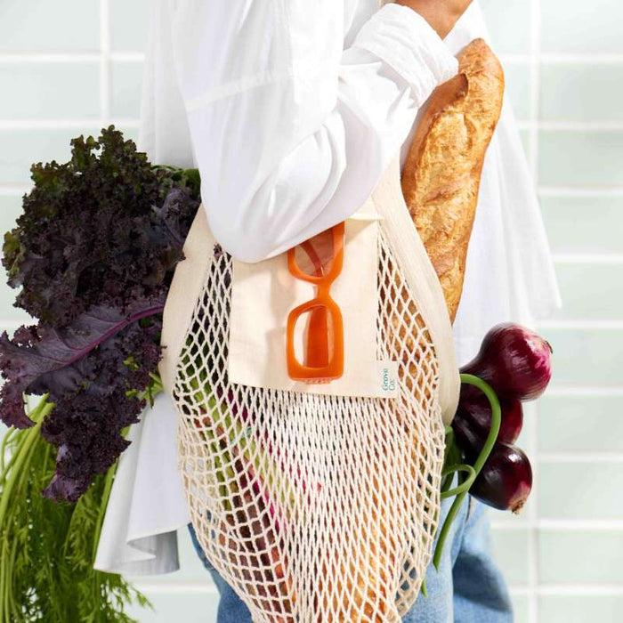 A person, dressed in a white shirt and blue jeans against a tiled background, embodies eco-friendly flair while carrying the stylish Grove Co. Mesh Farmers Market Tote filled with leafy greens, a baguette, carrots, and red onions; orange sunglasses rest on the bag.