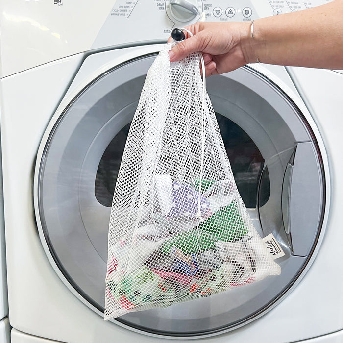 A person holds a Marleys Monsters Mesh Laundry Bag filled with colorful items, including UNpaper Towels, in front of an open pristine white washing machine with control buttons neatly arranged at the top.