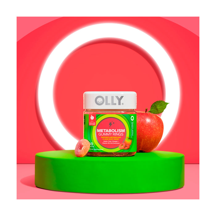 An OLLY Metabolism Gummy Rings jar, featuring Apple Cider Vinegar and Vitamin B12, rests on a green platform against a red backdrop. A red apple and a gummy accompany it, with an illuminated white circle in the background.