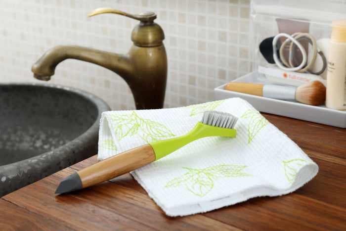 A bathroom sink with a brass faucet and gray basin includes a Full Circle Micro Manager Detail Brush with a green handle on a leaf-patterned cloth atop the wooden counter. Nearby, toiletries are neatly organized on a small tray.