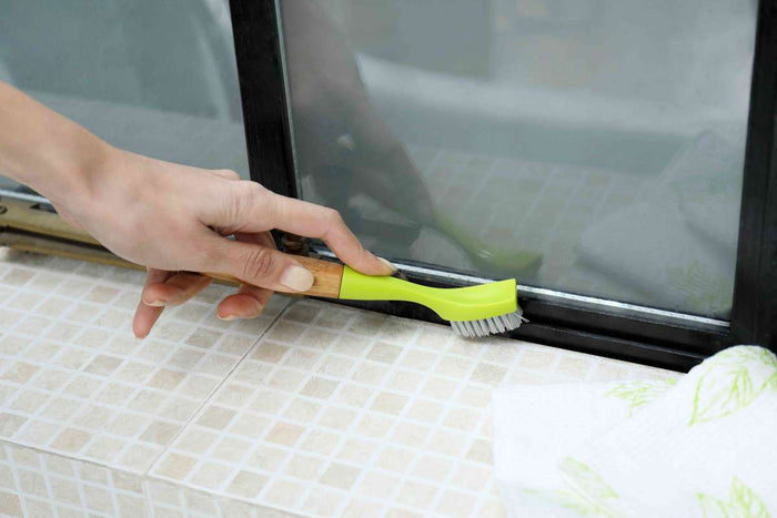 A meticulous person scrubs a sliding glass doors window track with Full Circles Micro Manager Detail Brush, featuring a green handle. The window is partially open, and the tiled surface beneath gleams as they embrace their inner Micro Manager.