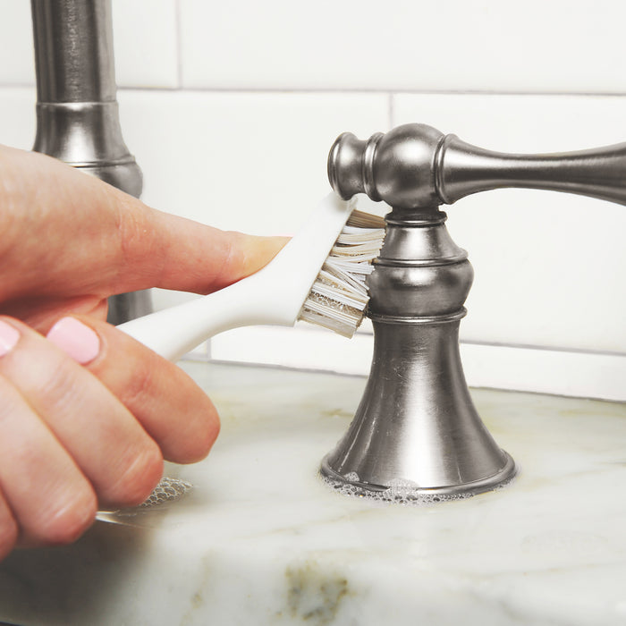 A hand uses the Full Circle Micro Manager Detail Brush to scrub around the base of a silver bathroom faucet on a marble countertop, with soap suds visible.