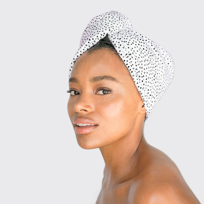 A woman wearing a luxuriously soft head wrap gazes at the camera, her clear skin glowing against a light background, highlighting how Kitschs Microfiber Hair Towel can gently care for your skin and hair.