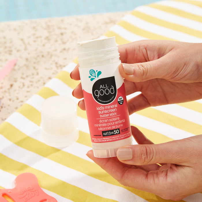 Close-up of hands holding the All Good Mineral Kids Sun Butter Stick - SPF 50 with organic ingredients. A striped towel and a glimpse of a pool in the background suggest a sunny outdoor setting ideal for kids sunscreen protection.