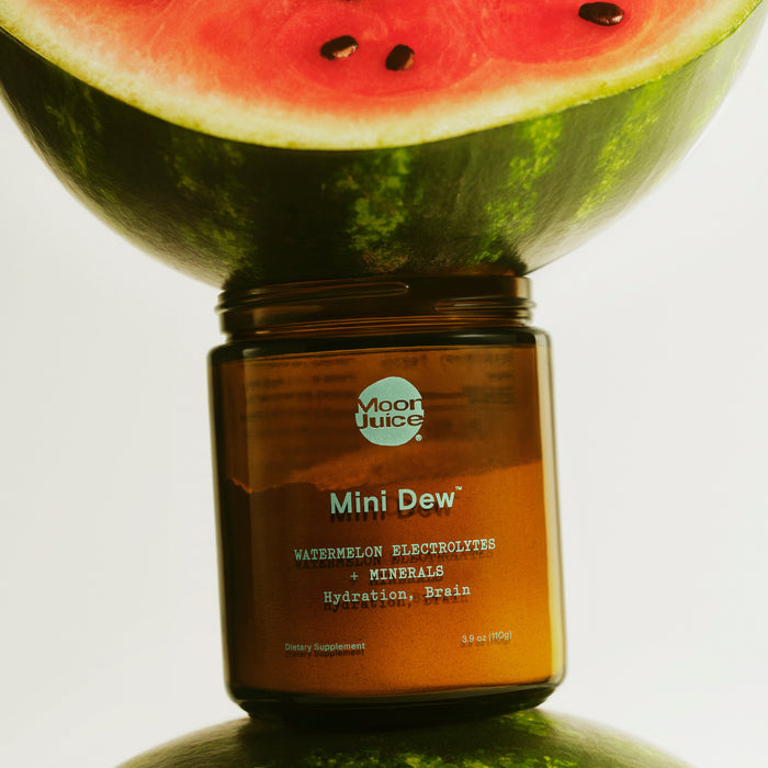 A jar of Moon Juices Mini Dew, a watermelon-flavored electrolyte supplement, sits between two watermelon halves. It is rich in essential minerals and promotes hydration and brain function. The watermelon halves show vibrant red flesh with small seeds.