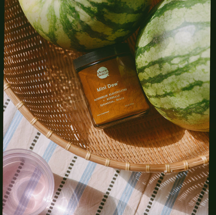 A wicker basket holds two large watermelons and a jar of Moon Juices Mini Dew melon preserves on a striped cloth, providing natural hydration and electrolytes. Nearby, a pink glass catches sunlight.