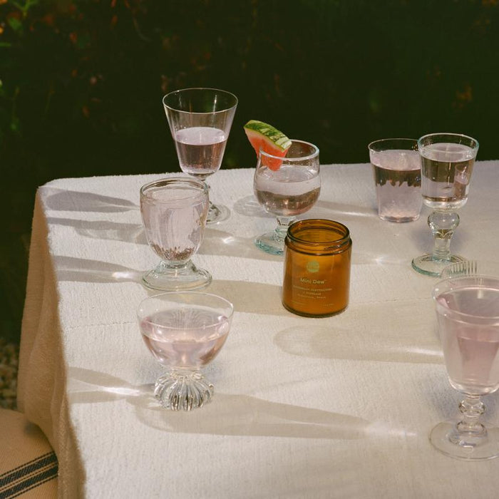 A table holds several glasses of pink-tinted water rich in electrolytes from Moon Juices Mini Dew. One glass is garnished with a watermelon slice for added hydration. A small amber candle adds a warm glow on the white-clothed table.