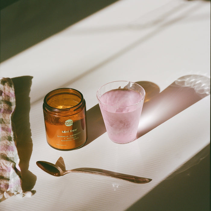 A jar labeled Mini Dew from Moon Juice sits beside a glass of pink electrolyte-rich liquid on a white table. A spoon is in front, with a checkered cloth to the left. Sunlight dances across, promising refreshing hydration and essential minerals.