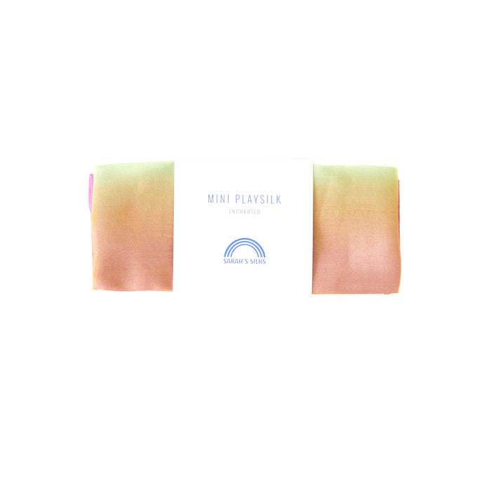 A Mini Playsilk by Sarahs Silks, made from 100% pure silk, features a pastel gradient of pink, peach, and green. It includes a label with MINI PLAY SILK ENCHANTED and a rainbow logo above SARAHS SILKS. Ideal for open-ended imaginative play.