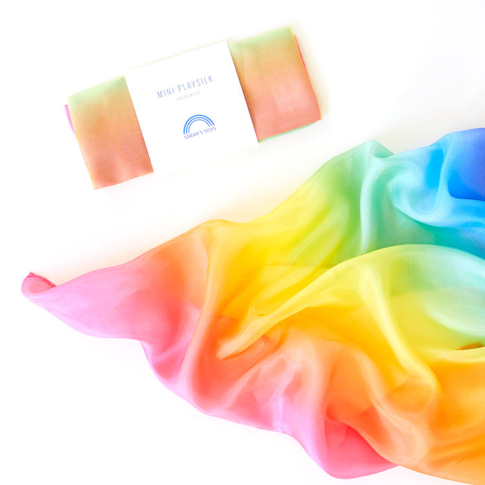 Sarahs Silks Mini Playsilk for Open-Ended, Imaginative Play features a colorful gradient of pink, orange, yellow, green, and blue. Made from 100% pure silk, its smooth and flowing texture is ideal for creative play. The packaged version is labeled Mini Playsilk.