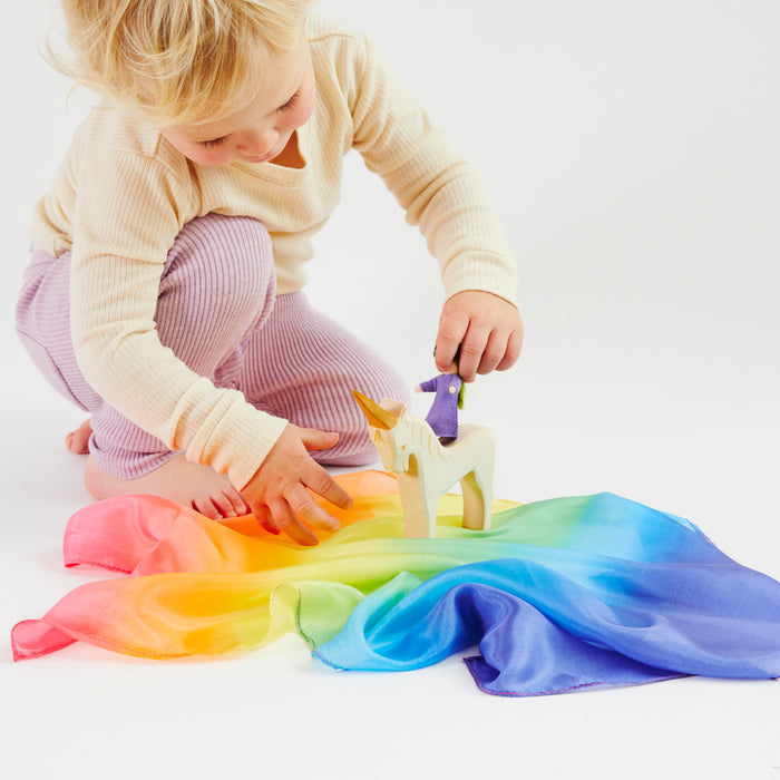A blond child engages in imaginative play with a wooden unicorn on a vibrant Mini Playsilk from Sarahs Silks, laid on the floor. Wearing a cream top and purple leggings, the child examines the toy against a plain white backdrop.