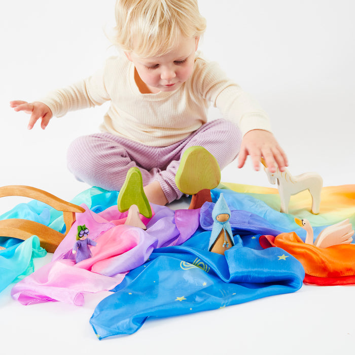 A blond child plays on the floor with colorful Mini Playsilk and imaginative wooden toys shaped like trees and animals. The vibrant scene, by Sarahs Silks, unfolds against a crisp white background.