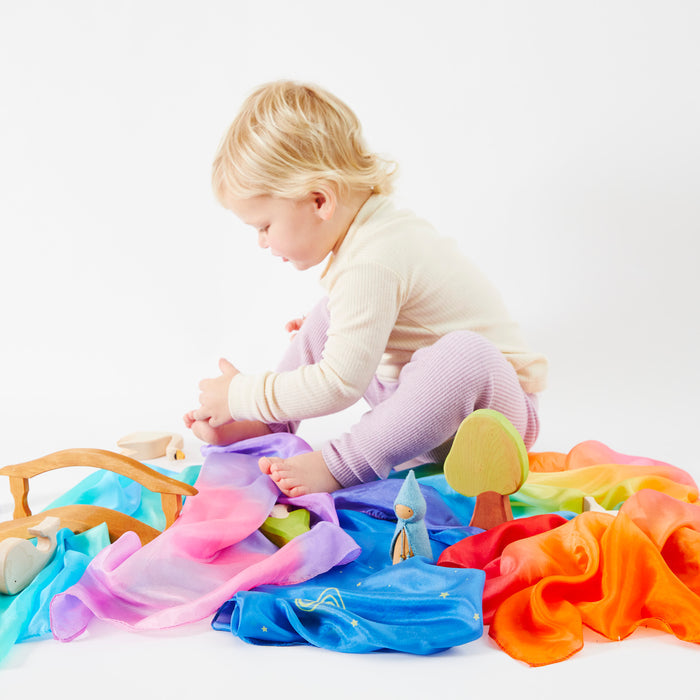 A toddler sits on the floor, surrounded by colorful Mini Playsilks from Sarahs Silks and wooden toys. Engrossed in open-ended play with a purple scarf, vibrant silks and toy figures are scattered around against a plain white background.