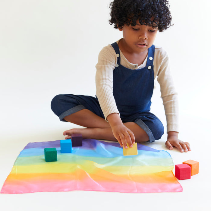 A curly-haired child sits against a white backdrop, absorbed in open-ended play. They wear a beige top with blue overalls and arrange colorful wooden blocks on Sarahs Silks Mini Playsilk for Open-Ended, Imaginative Play, which is spread out in rainbow hues before them.
