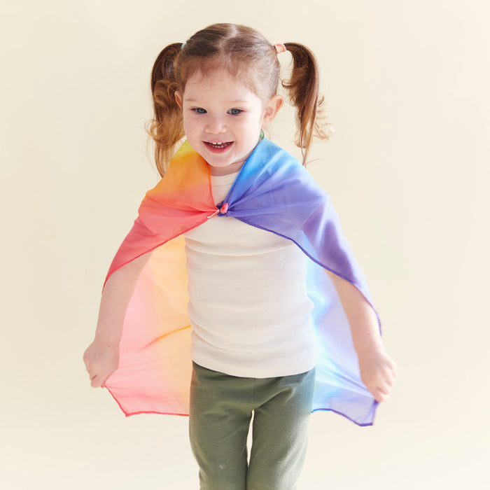 A young girl with pigtails smiles in a Mini Playsilk by Sarahs Silks, draped in a yellow-to-purple gradient cape. Her white shirt and green pants complete the look, celebrating imaginative play against a light background.